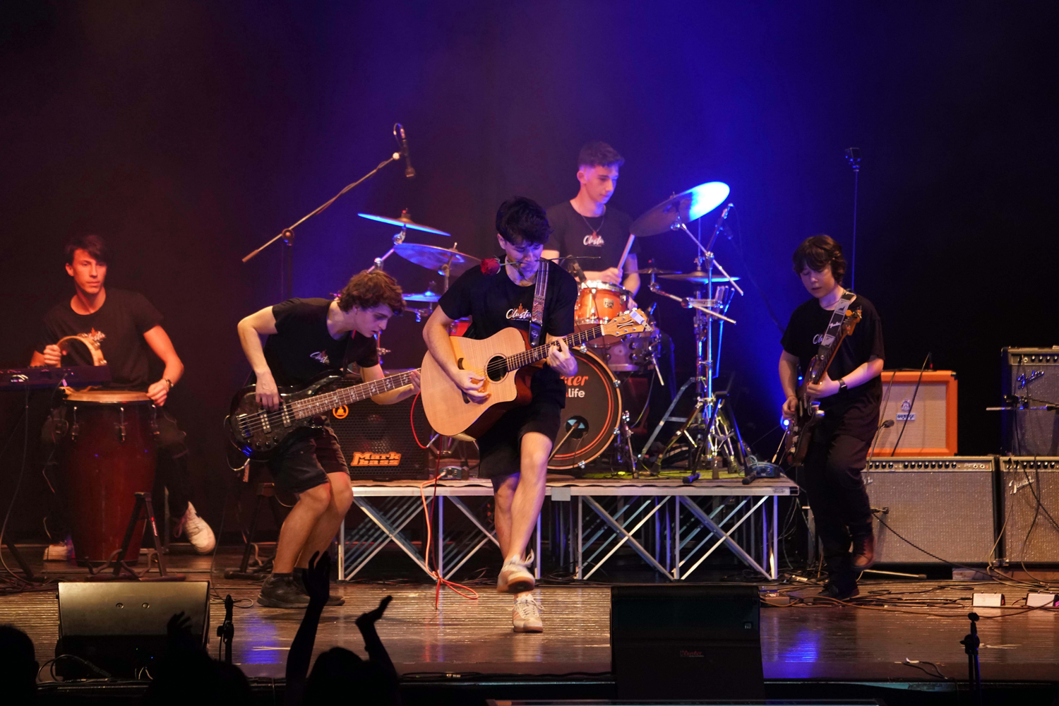 Concerto live con studenti della Scuola e Accademia di Musica Cluster: chitarrista acustico, bassista e batterista si esibiscono su palco illuminato, in un evento musicale scolastico
