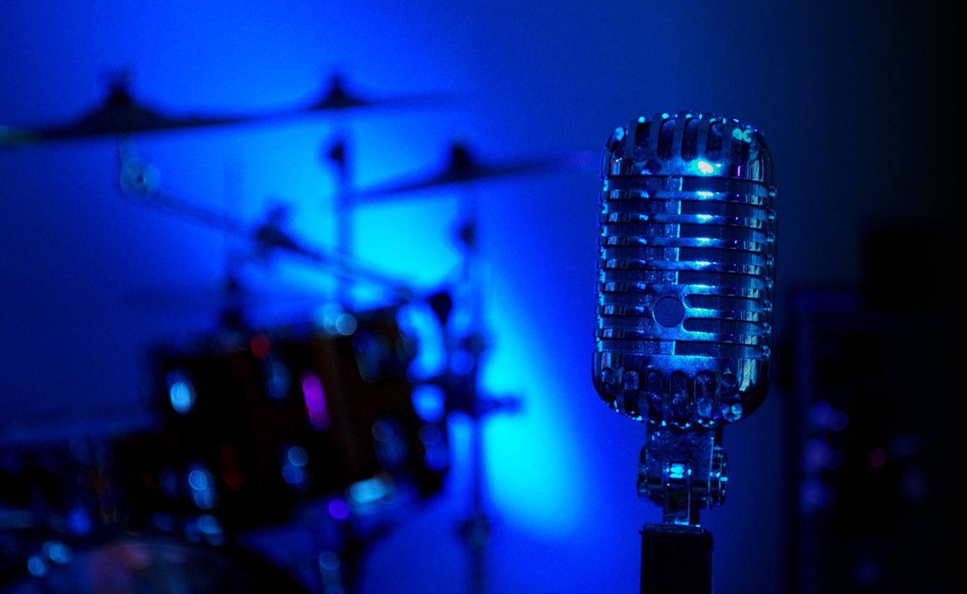 Microfono da studio illuminato da luce blu con batteria sullo sfondo, simbolo delle lezioni di canto moderno alla Scuola e Accademia di Musica Cluster